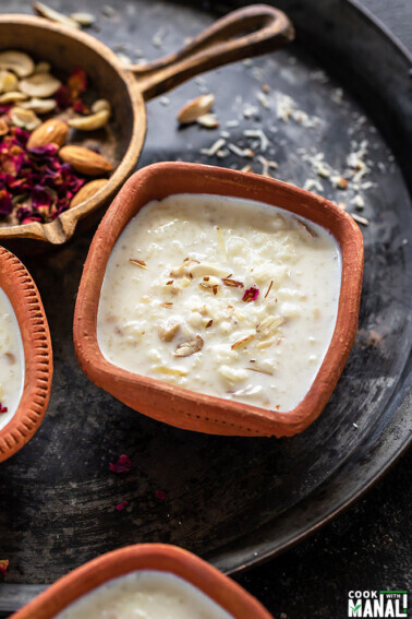 Rice Kheer served in a clay bowl and garnished with rose petals and a golden cast iron pan with nuts and rose petals in the background
