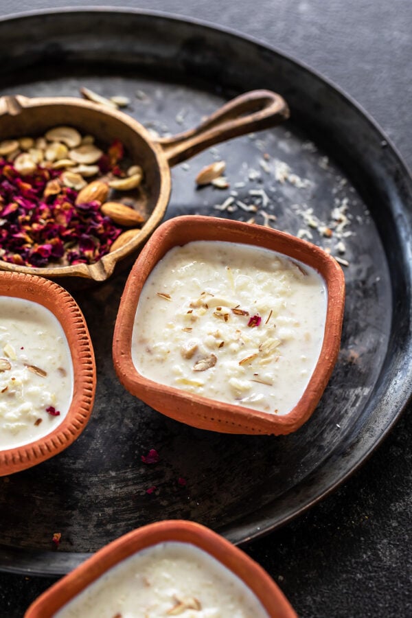 Rice Kheer served in a clay bowl and garnished with rose petals and a golden cast iron pan with nuts and rose petals in the background