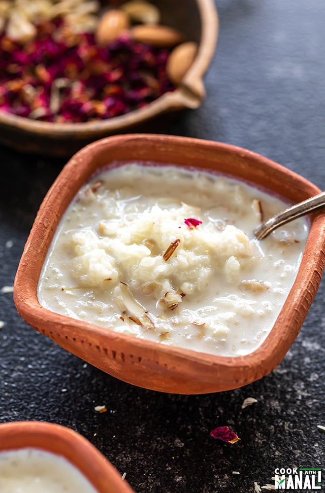 kheer served in a clay bowl with a spoon and a pan of nuts and rose petals in the background