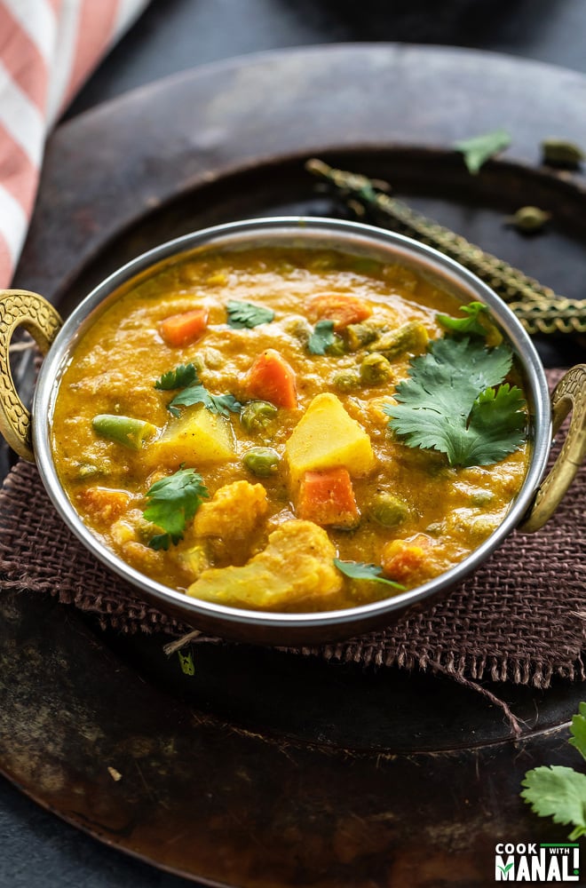 veg kurma served in a copper kadai with a striped kitchen towel placed on the side and golden spoon on the other side