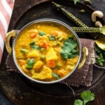 overhead shot of veg kurma served in a copper kadai with a kitchen towel placed on one side and two golden color antique spoon on the other side