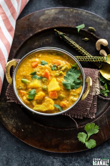 overhead shot of veg kurma served in a copper kadai with a kitchen towel placed on one side and two golden color antique spoon on the other side