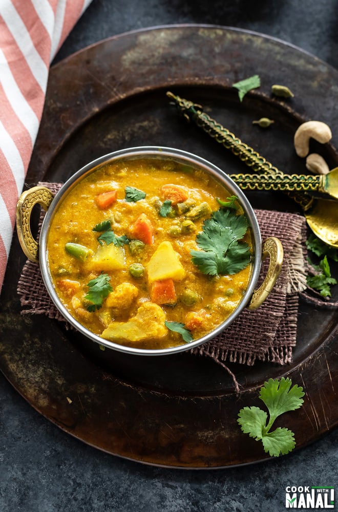 overhead shot of veg kurma served in a copper kadai with a kitchen towel placed on one side and two golden color antique spoon on the other side