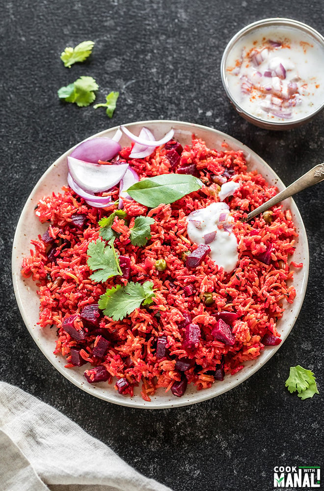 overhead shot of beetroot pulao in a white plate with a spoon and a small bowl of yogurt in the background