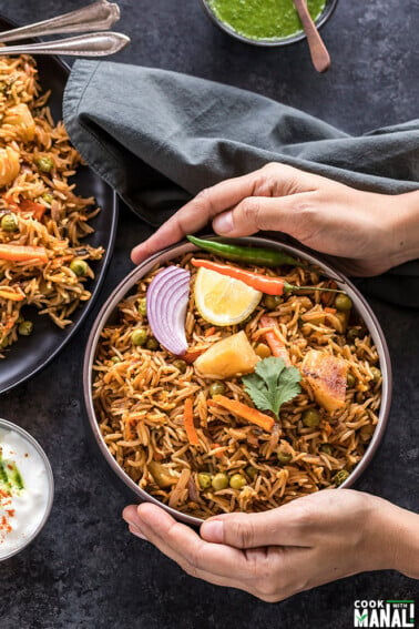 vegetable biryani in a black bowl with a bowl of yogurt on the side