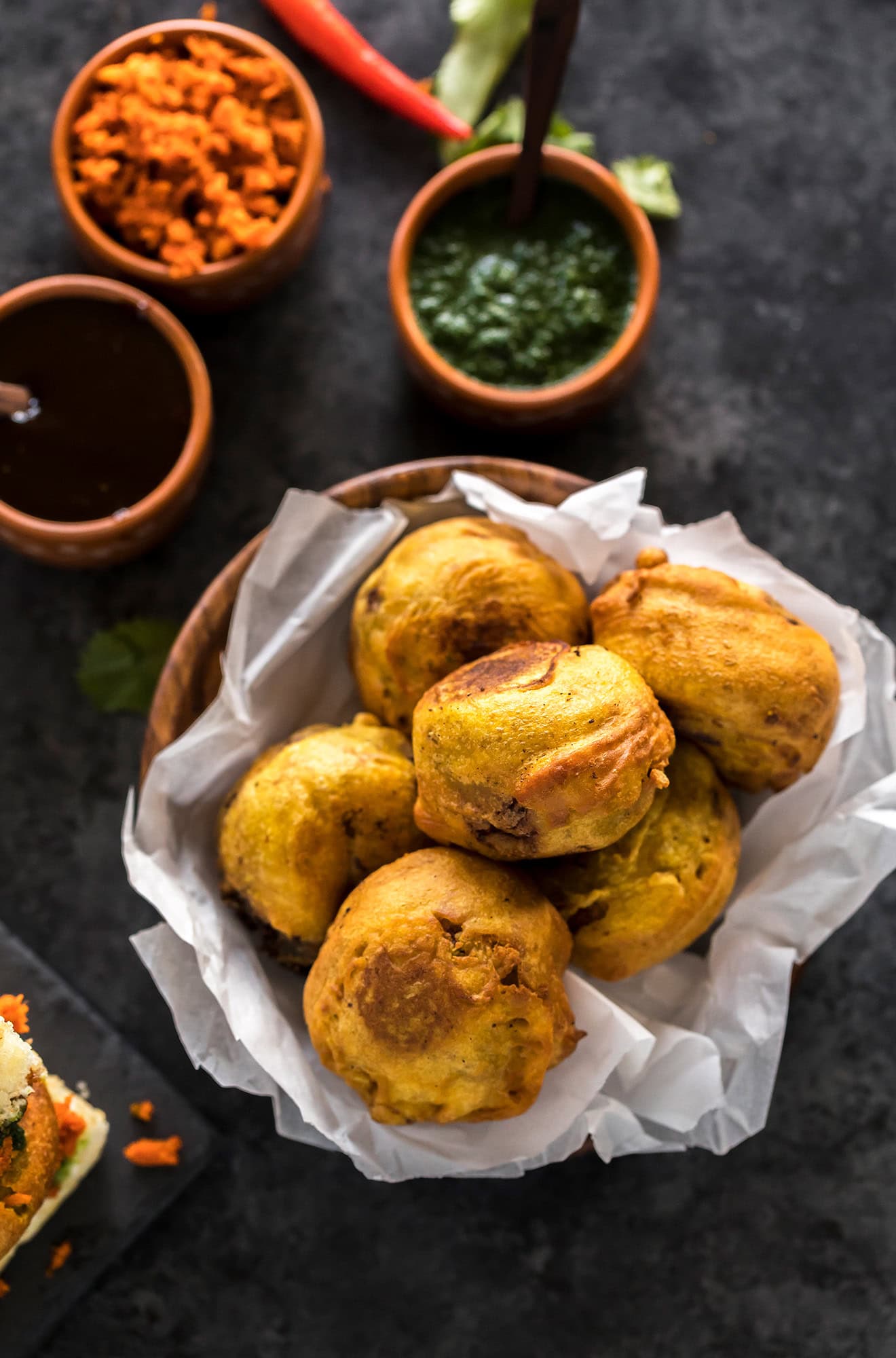 batata vada served in a wooden bowl lined with parchment paper with chutney bowls on the back