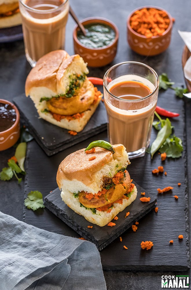 Mumbai Vada Pav placed on a black coaster with glass of chai and bowls of chutney in the background