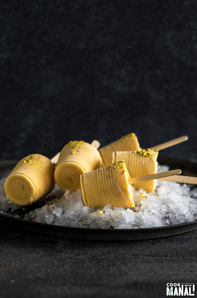 mango kulfi popsicles on top of crushed ice on a black plate