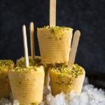 mango kulfi popsicles on top of crushed ice on a black plate