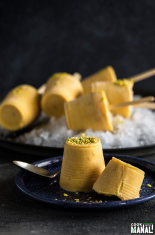 mango kulfi garnished with pisachios and served on a blue plate with more mango kulfi popsicles in the background