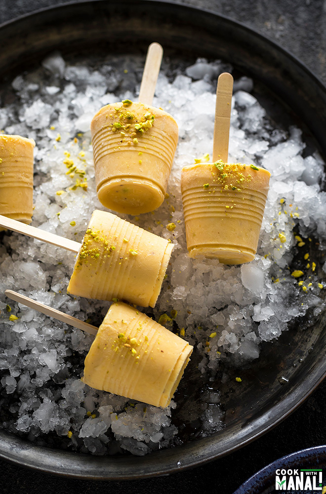 mango kulfi popsicles on top of crushed ice on a black plate