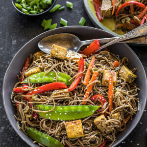 vegan soba noodles with carrots, snow peas, tofu, red pepper in a large grey bowl with a grey napkin on the side