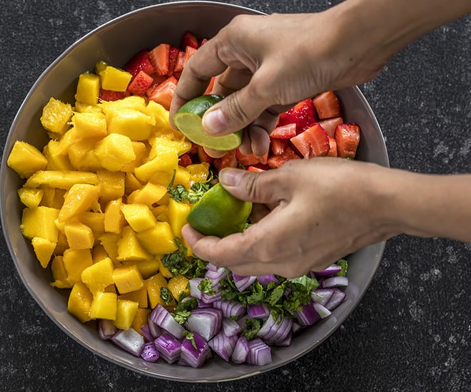 a pair of hand squeezing lime juice into a bowl of strawberry mango salsa