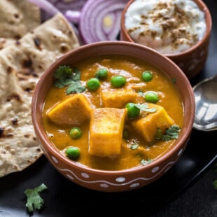 matar paneer in a clay bowl with a bowl of yogurt on the side and some rotis in the back