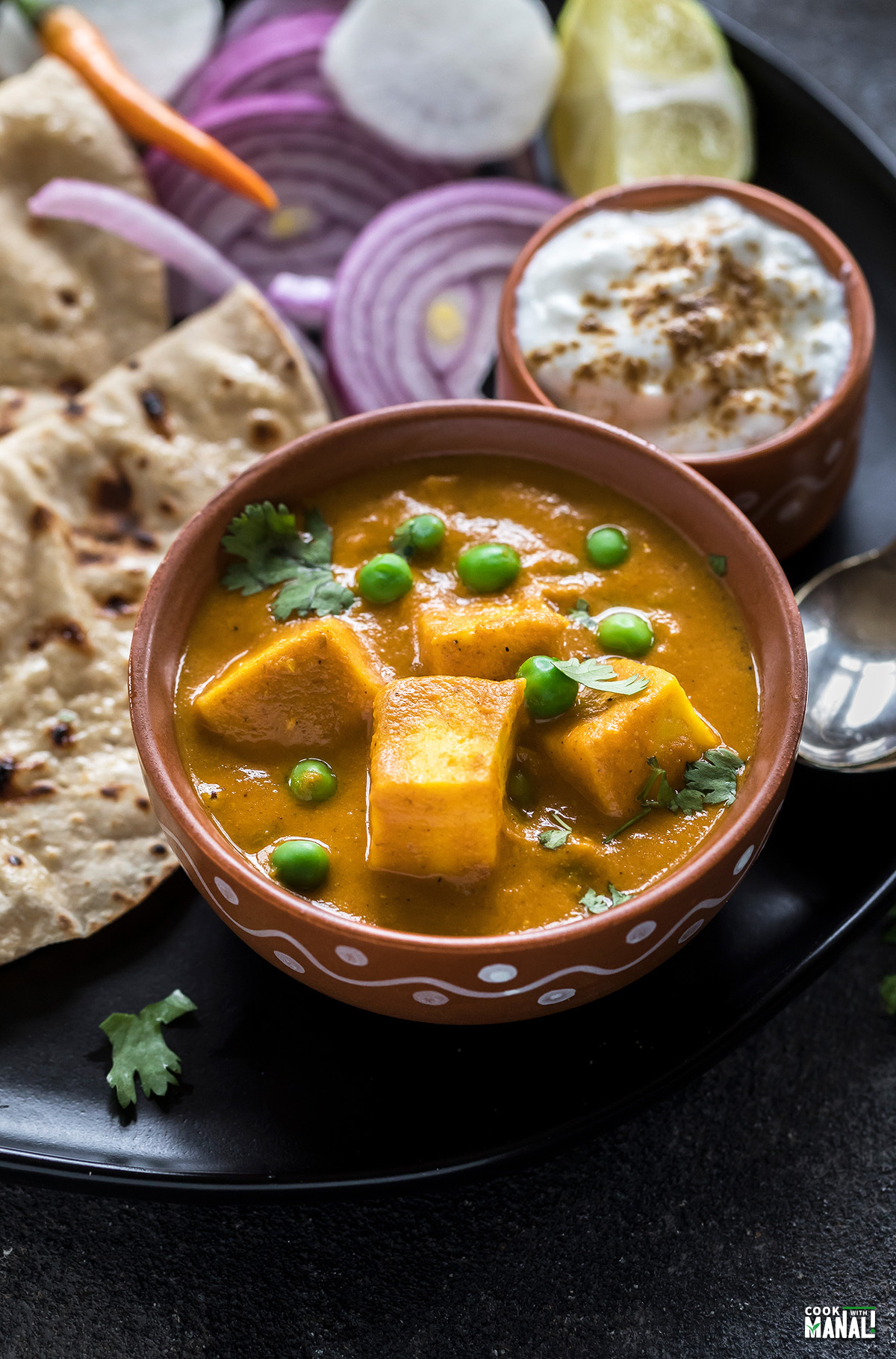 matar paneer in a clay bowl with a bowl of yogurt on the side and some rotis in the back