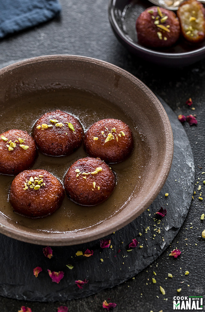 five pieces of gulab jamun in a brown round place with dried rose petals sprinkled all around