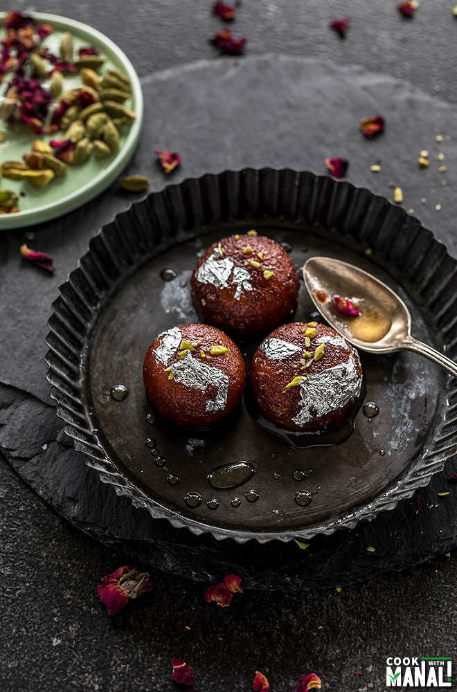 three pieces of gulab jamun in a round black plate with cardamom and dried rose petals in the background