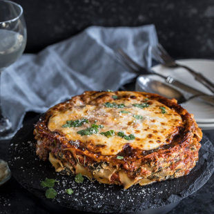 spinach mushroom lasagna on a round black serving board with a napkin and glass in the background