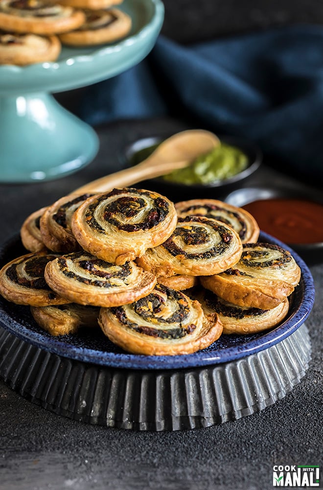 spinach & sun-dried tomato puff pastry pinwheels stacked on a blue plate with black bowls filled with chutney & ketchup in the background