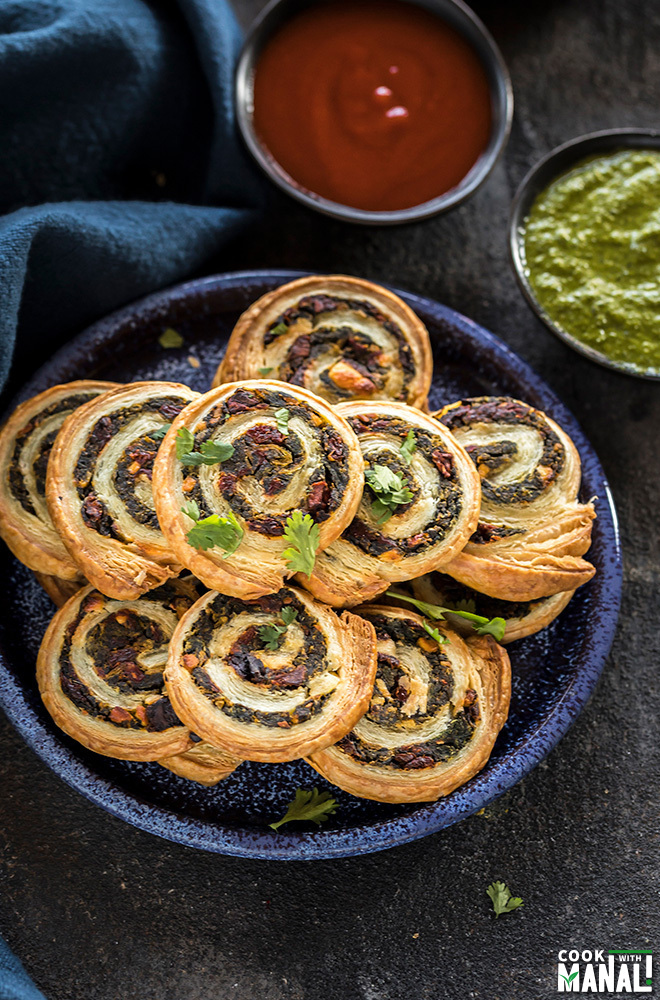 spinach & sun-dried tomato puff pastry pinwheels stacked on a blue plate with black bowls filled with chutney & ketchup in the background