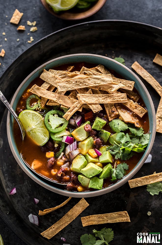 bowl of vegetarian tortilla soup with a spoon and topped with avocado, tortilla strips, cilantro, lime wedge with more garnishes on the side