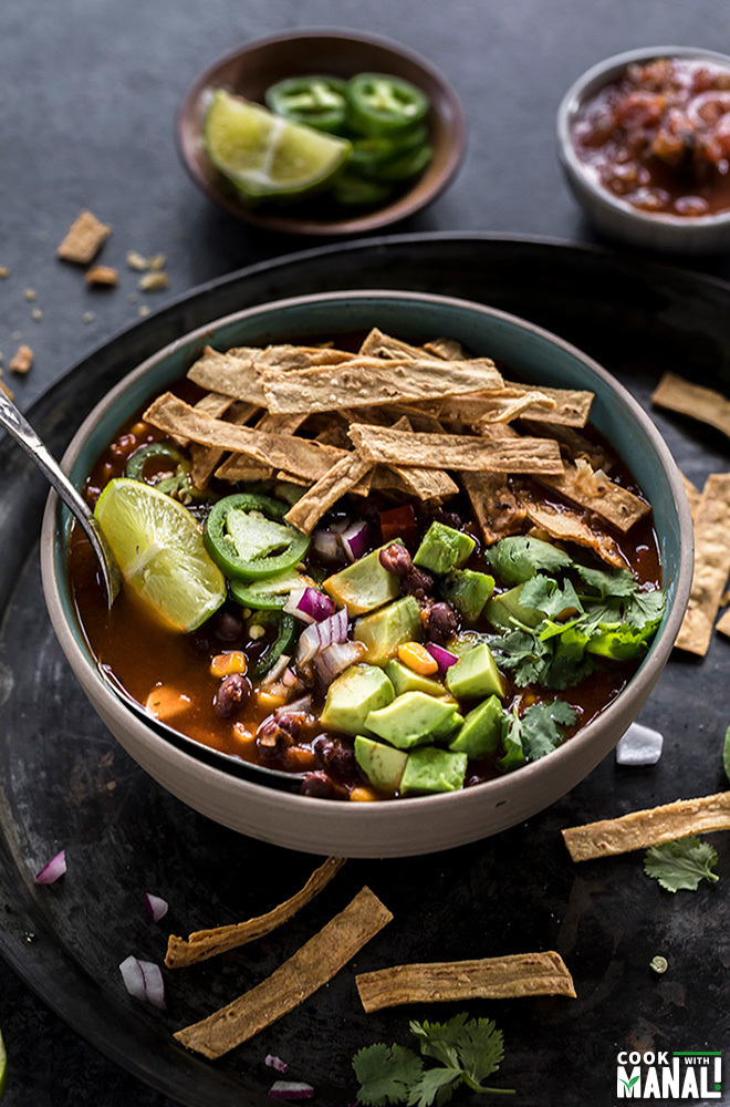 bowl of vegetarian tortilla soup with a spoon and topped with avocado, tortilla strips, cilantro, lime wedge with more garnishes on the side