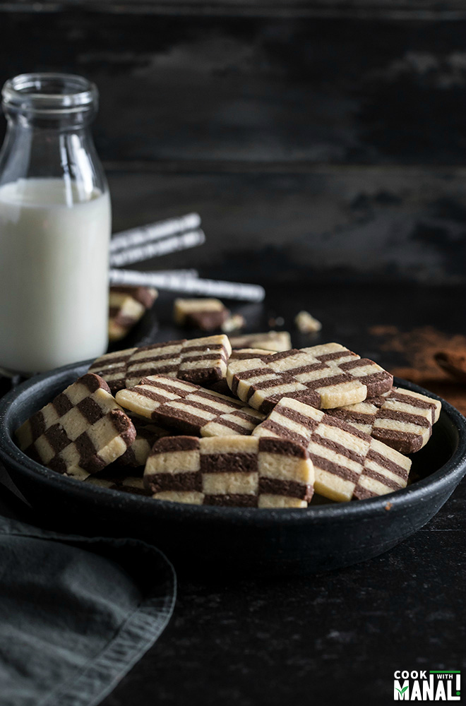 eggless checkerboard cookies in a black bowl with a bottle of milk and straws in the background