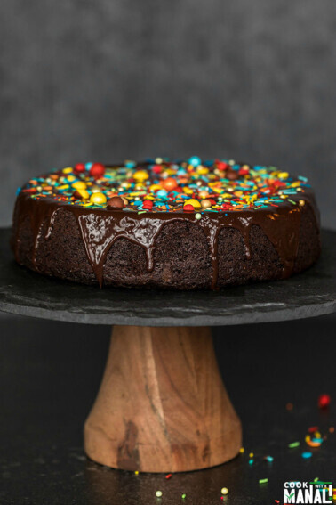 eggless chocolate cake on a black cake stand, covered with chocolate ganache and sprinkles