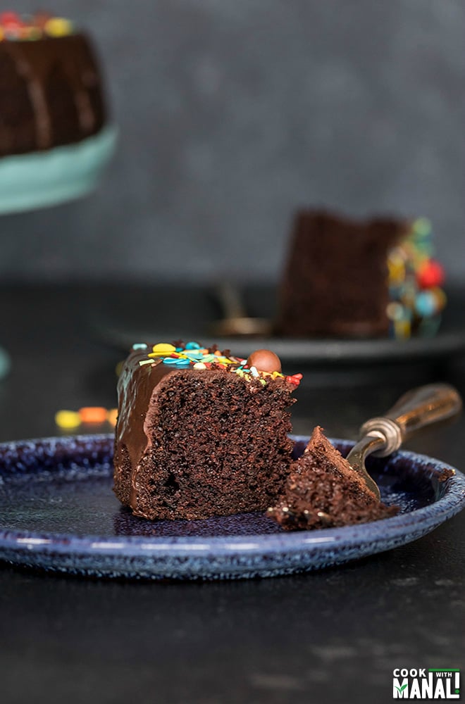 slice of eggless chocolate cake in a blue plate with a fork on the side with another plate and cake stand in the background