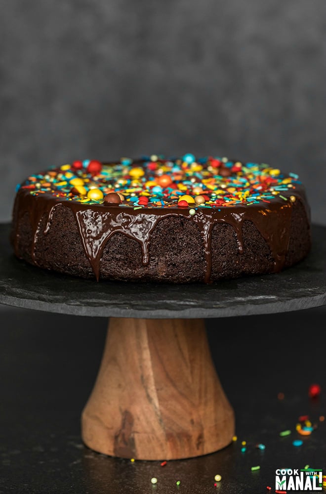 eggless chocolate cake on a black cake stand, covered with chocolate ganache and sprinkles