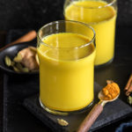 haldi doodh aka golden milk in a glass with another glass in the background and a spoon with turmeric on the side