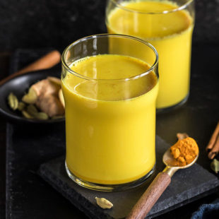 haldi doodh aka golden milk in a glass with another glass in the background and a spoon with turmeric on the side