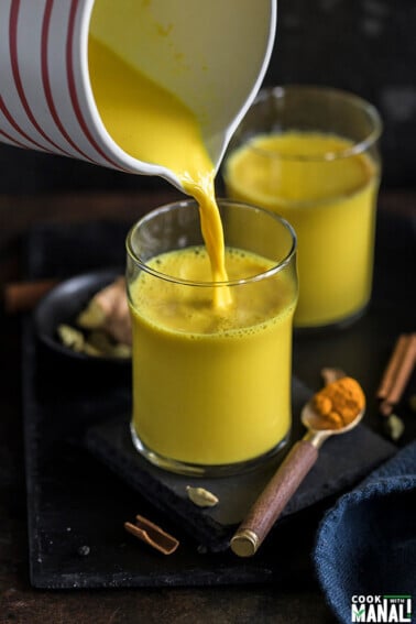 golden milk being poured from a jug into a glass with another glass in the background and a spoon full of turmeric on the side