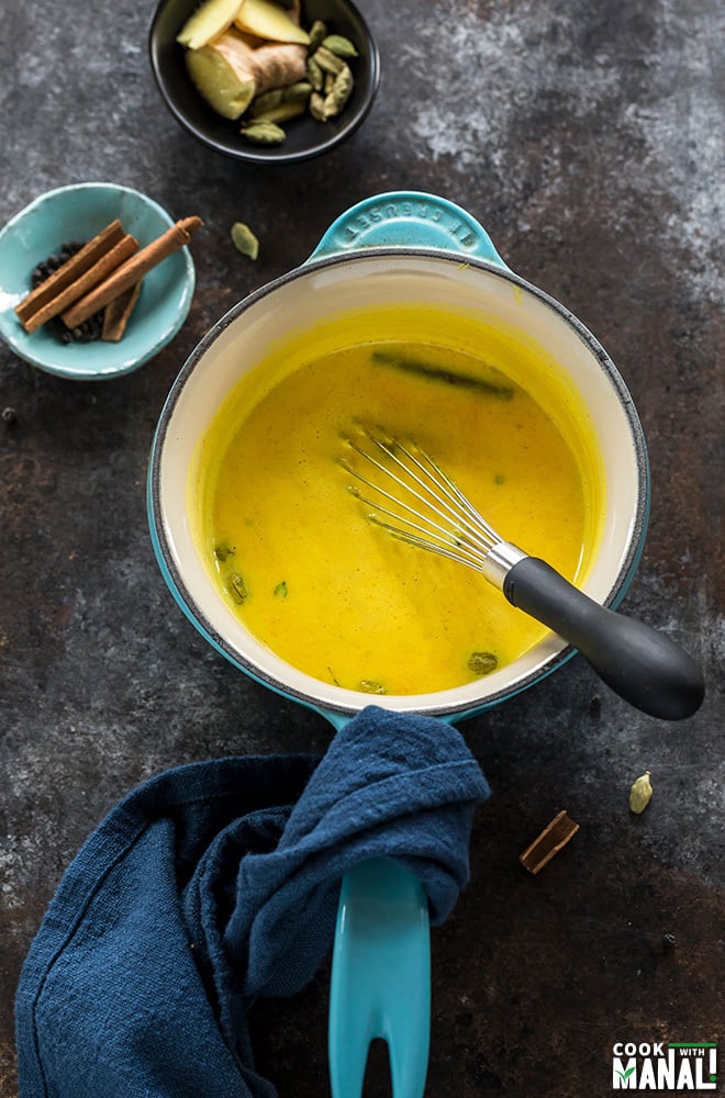 golden milk in a blue pot with a whisk with small bowls of spices on the side