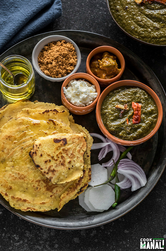 a thali with sarson ka saag in a bowl along with makki ki roti and several other bowls of pickle, butter, jaggery and ghee