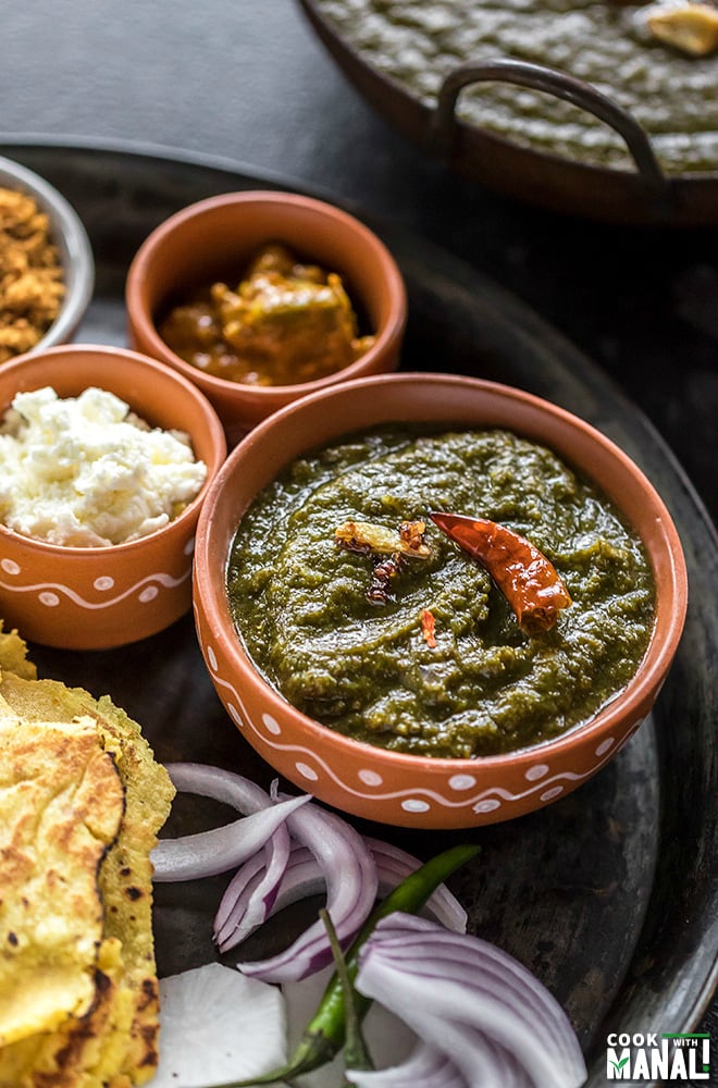 sarson ka saag in a small clay bowl with several other bowls filled with pickle, butter, jaggery