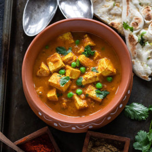 tofu matar served in a round clay pot with naan and spoons in the background and spice boxes on the side