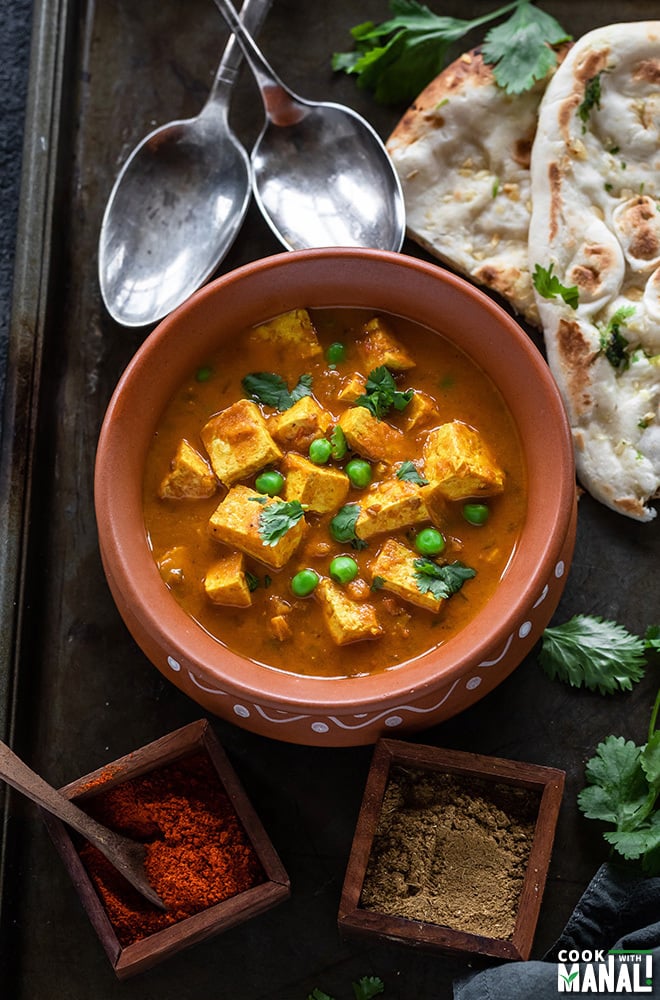 tofu matar served in a round clay pot with naan and spoons in the background and spice boxes on the side