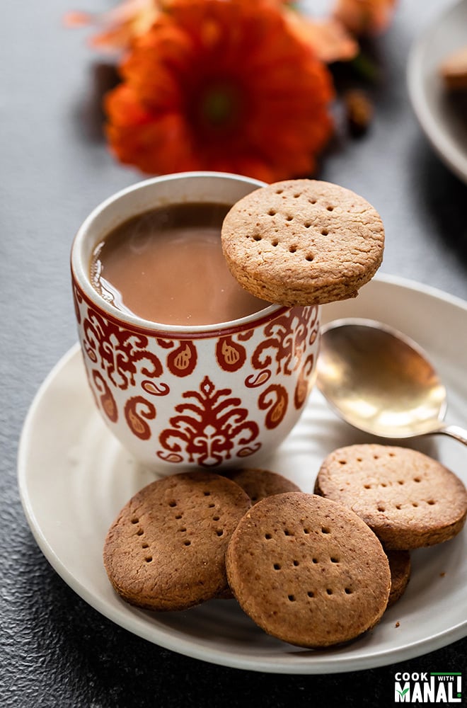 jaggery cookie place on the side of a cup of chai with more cookies in the plate below