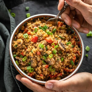 a hand holding a bowl of mushroom cauliflower fried rice from one side and digging into the bowl with a spoon from the other side