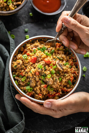 a hand holding a bowl of mushroom cauliflower fried rice from one side and digging into the bowl with a spoon from the other side