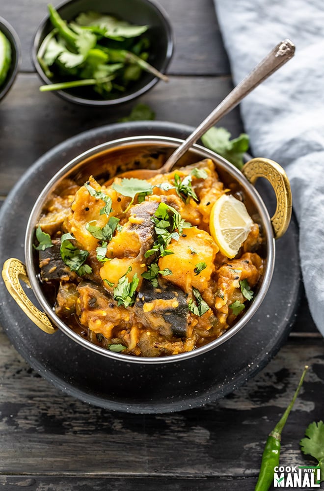 aloo baingan in a copper kadai with a spoon with a napkin on the side and a bowl of chilies in the background