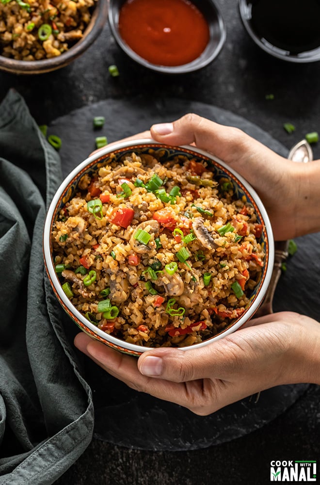 pair of hands holding a bowl of mushroom cauliflower fried rice with napkin on the side and sauce bowls in the back