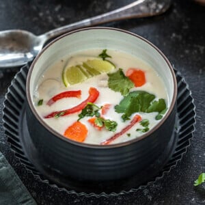 vegan tom kha gai in a black bowl placed in a black rimmed plate with a spoon in the background and some cilantro on the side