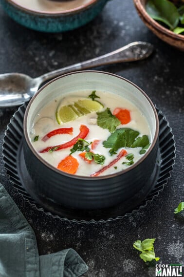 vegan tom kha gai in a black bowl placed in a black rimmed plate with a spoon in the background and some cilantro on the side