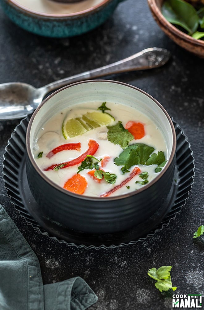 vegan tom kha gai in a black bowl placed in a black rimmed plate with a spoon in the background and some cilantro on the side