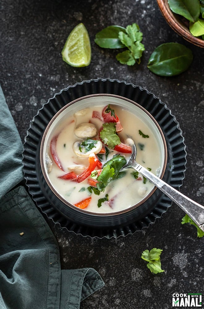 overhead shot of vegan tom kha gai soup in a black bowl with a spoon and some garnished like lime wedge and cilantro on the sides