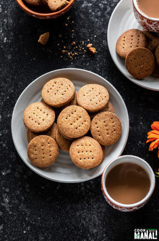 whole wheat jaggery cookies in a white plate with cups of chai on the side