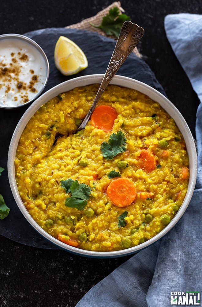 bowl of quinoa khichdi with a spoon with a small bowl of yogurt and lemon slice in the background