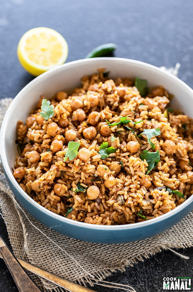 brown rice chana pulao served in a blue bowl, garnished with cilantro and a cut lemon and green chili in the background